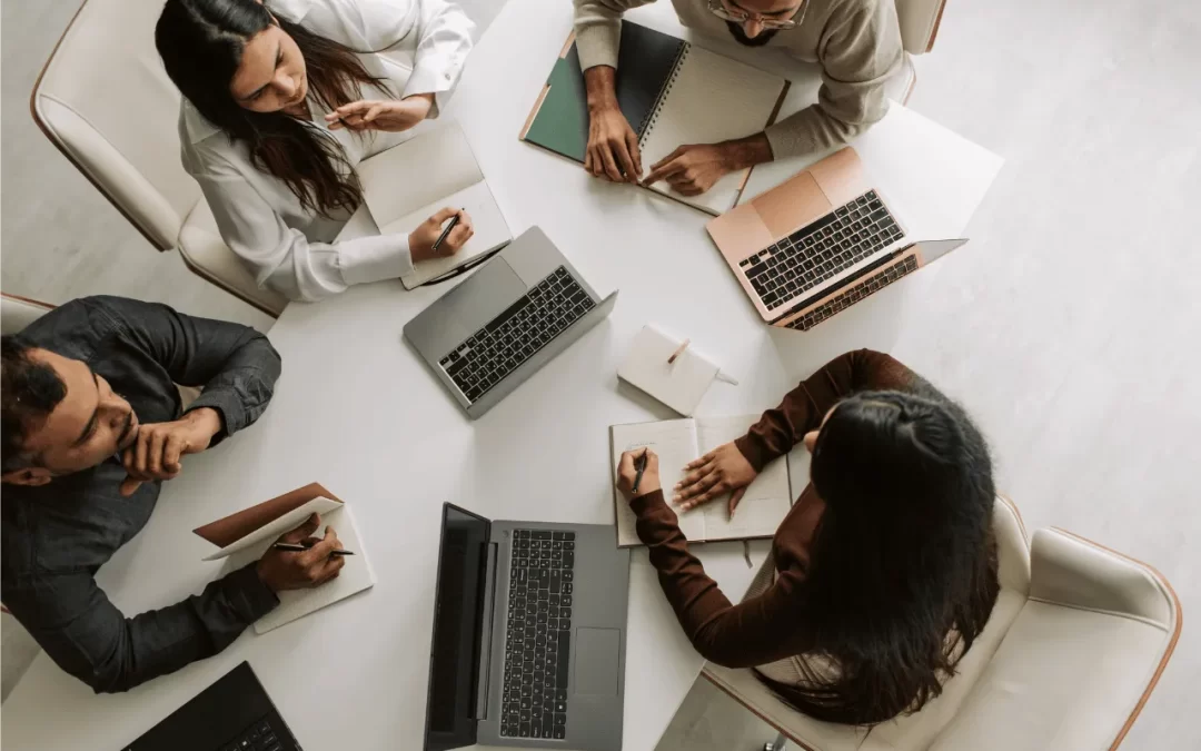Image of a marketing agency in a brainstorm. Photo features four individuals around a table with notebooks and laptops.